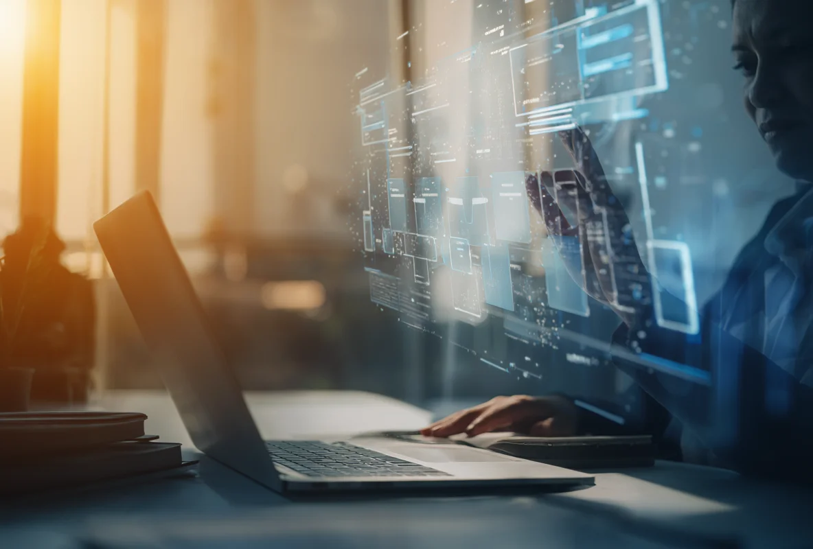 A person at a desk works on a laptop while touching a glowing holographic AI data dashboard, bathed in warm window light.