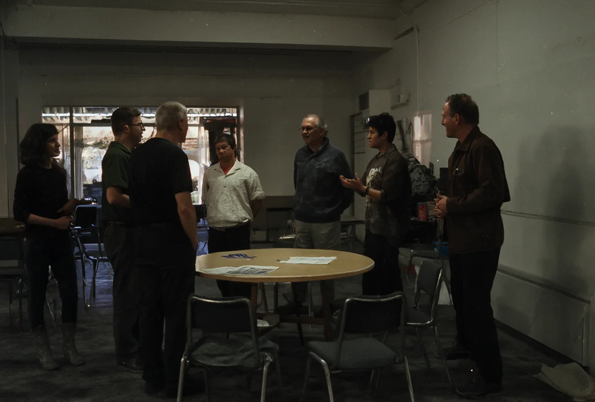 Six adults stand in a circle around a round table covered with papers, having a discussion in a dimly lit room.